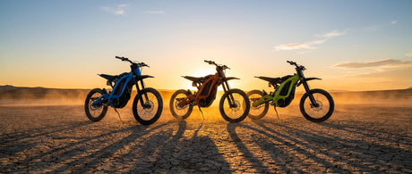 Three electric dirt bikes lined up side by side on a cracked desert hardpan under golden hour light, casting long dramatic shadows.